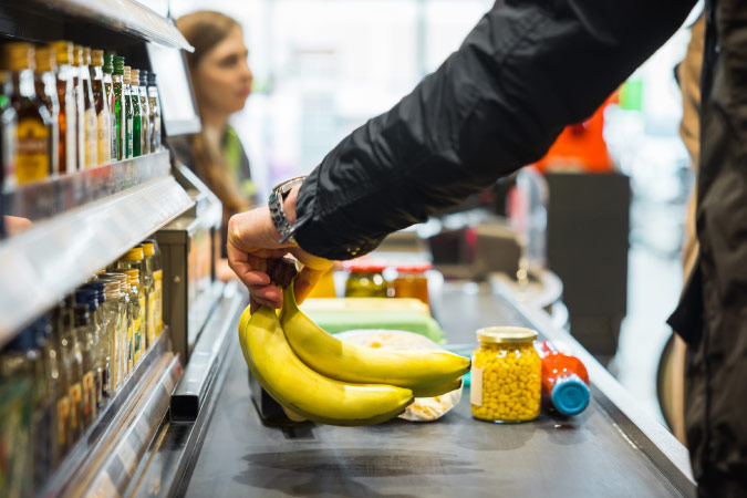 Closeup of a man’s arm setting a bunch of bananas on a conveyor belt at the grocery store checkout counter.