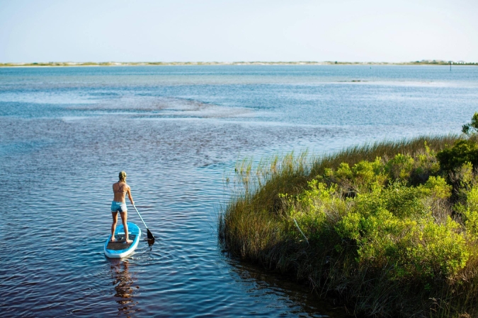 A paddleboarder is gliding through calm coastal waters and marshland, one of many relaxing things to do in Pensacola, Florida.