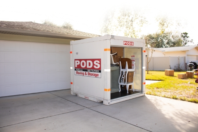 A PODS container filled with furniture sits in a driveway with moving boxes scattered in the front lawn.