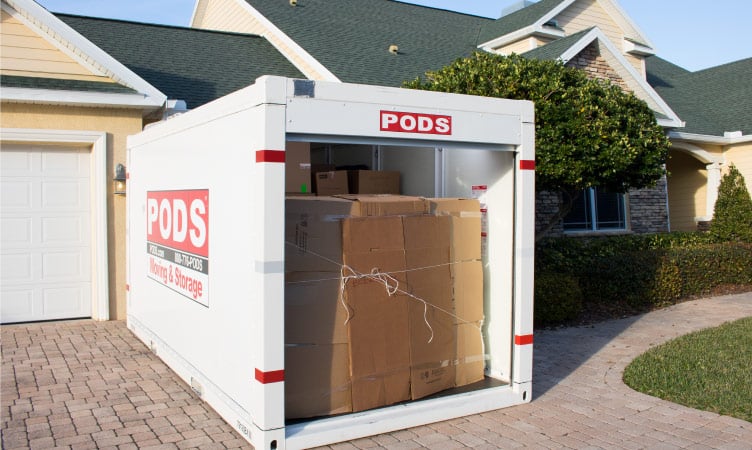A PODS moving container sits in the driveway of a tan-colored home, loaded with moving boxes that are secured with rope.