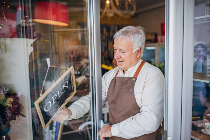A senior man with a canvas apron turns a sign to “OPEN” in the window of his boutique flower shop, like those you’d find while living in Fort Collins, Colorado.