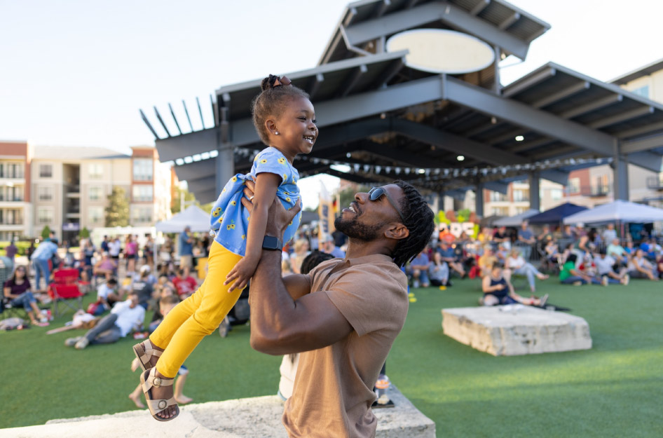 A father is holding his smiling young daughter up in his arms at a local event in Pearland, Texas.