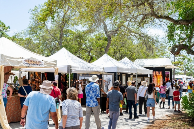 Dozens of locals living in Melbourne, FL, walk along a tent-lined path of vendors at the Melbourne Art Festival, showcasing various artists on a sunny day.