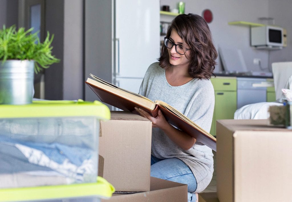 A young woman kneels on the floor as she looks through a photo album. Thre are boxes scattered around the kitchen. 