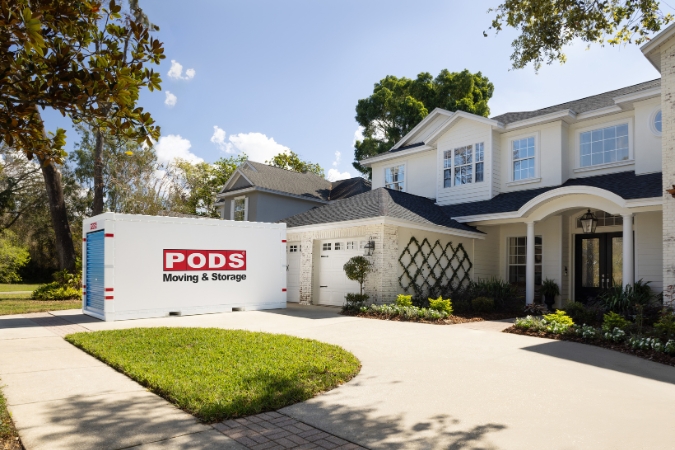 A PODS portable storage container sits in the driveway of a two-story home with a manicured lawn and landscaped front entry.