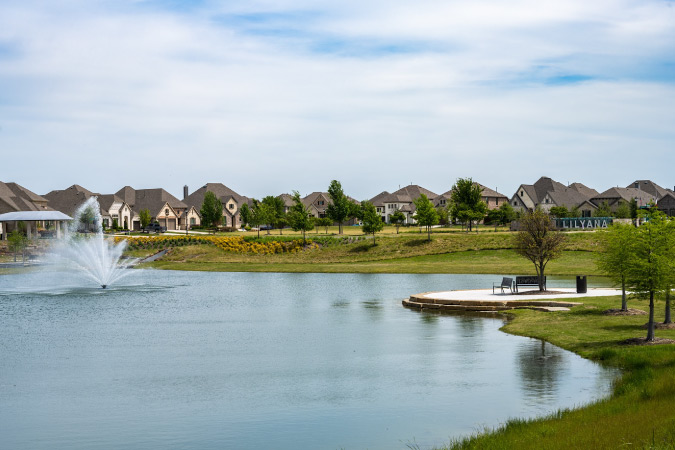 View over a tranquil pond in Lilyana — one of the best Celina, TX, neighborhoods — featuring a water fountain and surrounded by new construction, single-family homes.