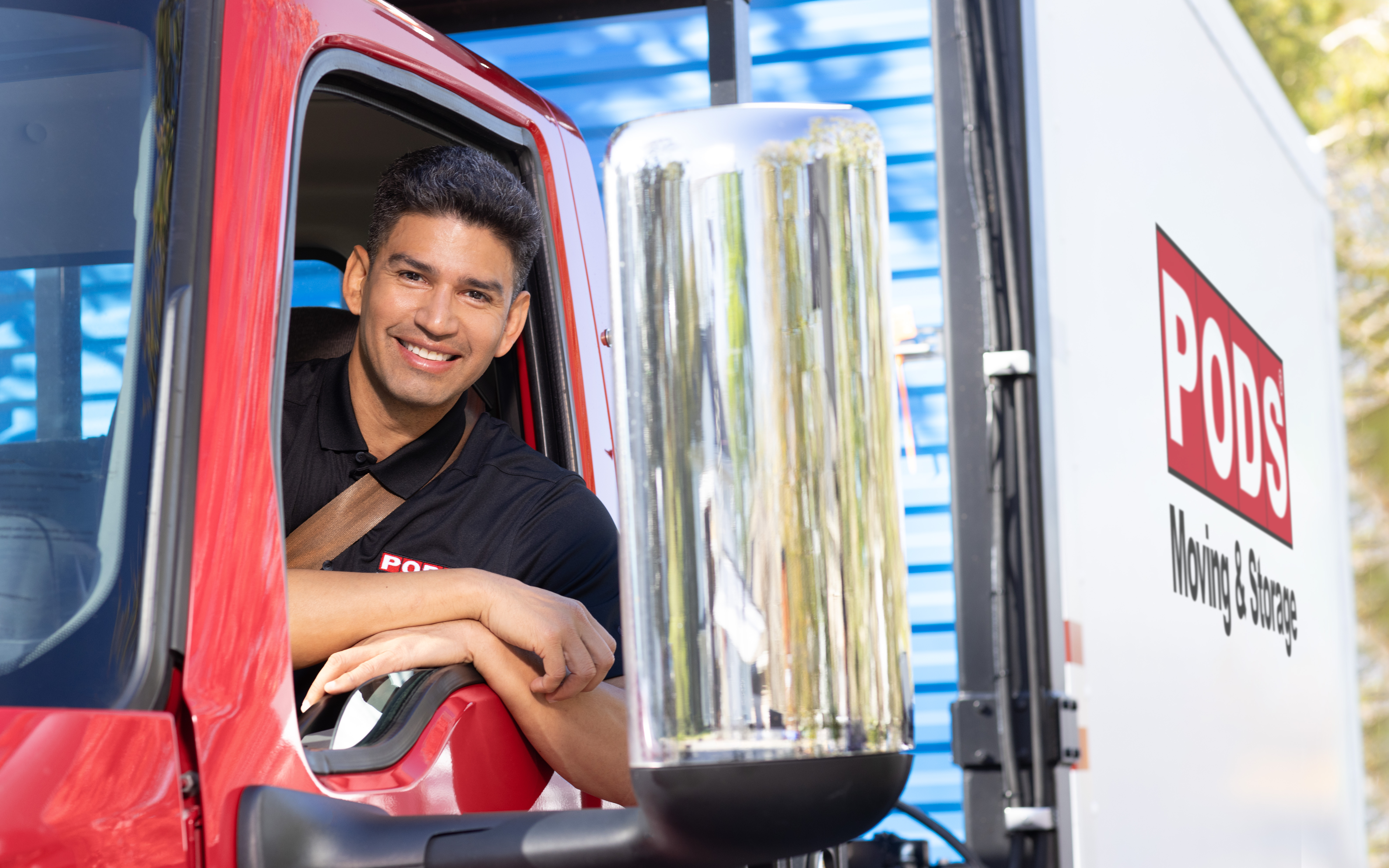 A PODS driver is smiling from the cab of his truck. He’s parked in front of a residential home, and there’s a PODS portable moving and storage container on the back of the truck.