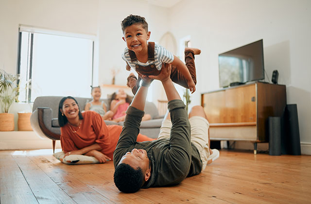A father lifts his son in the air as he lies on his back. Mom and daughters look on from a pillow on the floor and the sofa, respectively.