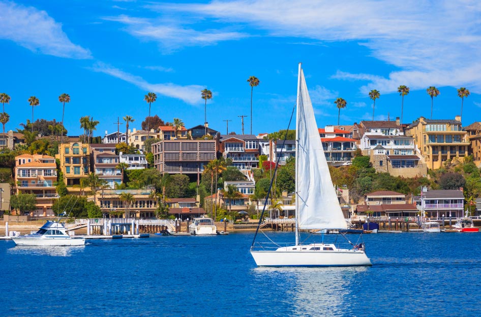 A sunny summer day at Newport Bay in Newport Beach, California. The waterfront is packed with condos and commercial buildings, as a small sailboat glides past the coast.