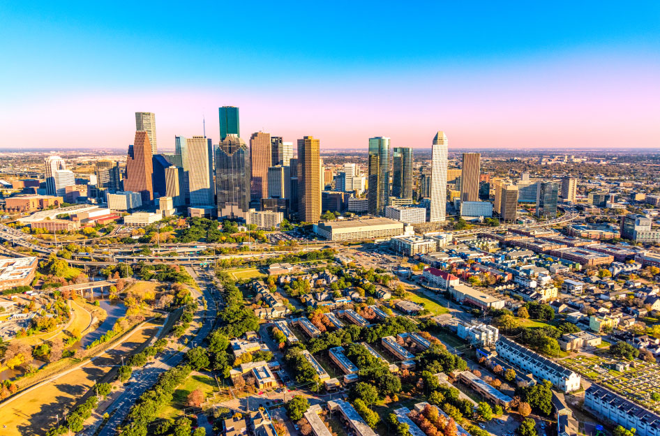 Sunny aerial view of the Houston skyline and some of the best neighborhoods in Houston, featuring a pink and blue sky and rows of lush trees lining the streets.