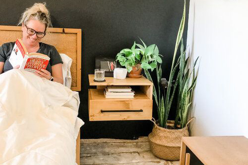 a woman with blonde hair reds in her bed with a light wooden nightstand sits off to the side. 