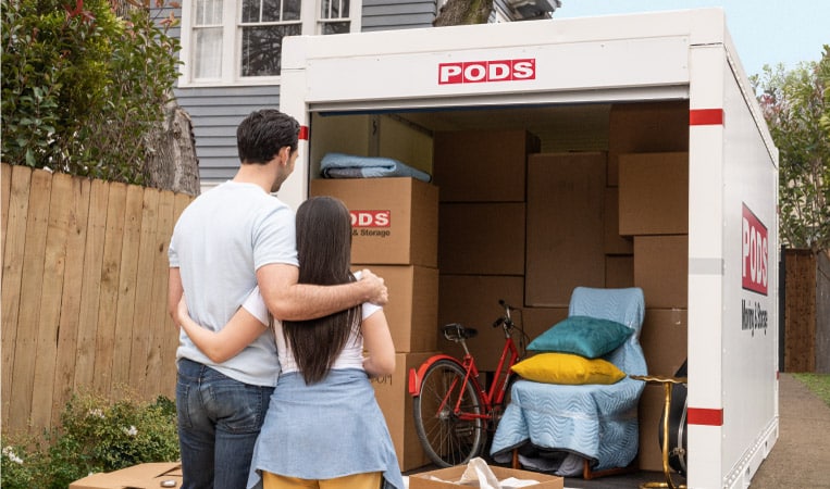 A couple stands with their arms around each other's shoulders, looking on at their partially loaded PODS moving and storage container. They’re in the process of moving to Dallas, TX.