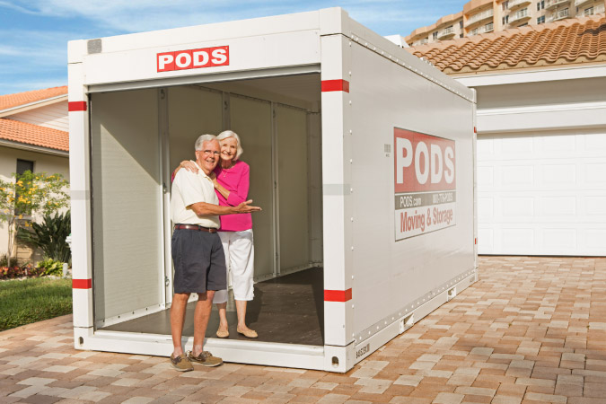 A senior couple smiles at the entrance of an empty PODS portable container in their driveway, which is ready to be loaded with items during estate sale planning.
