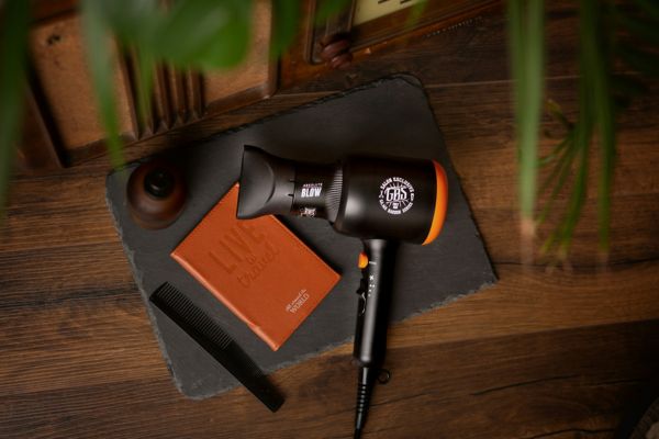 A black and orange hair dryer left on top of a wooden table right next to an orange notebook and a black comb.