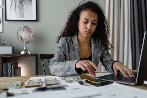 A woman in an office setting calculates her online store profitability with the help of a calculator and a laptop.
