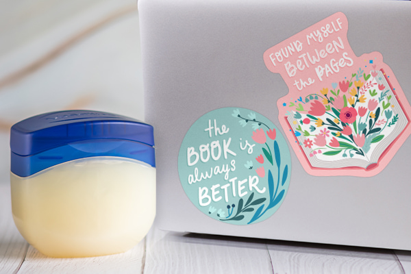 A laptop with stickers on its cover and a jar of an oil-based product right next to it